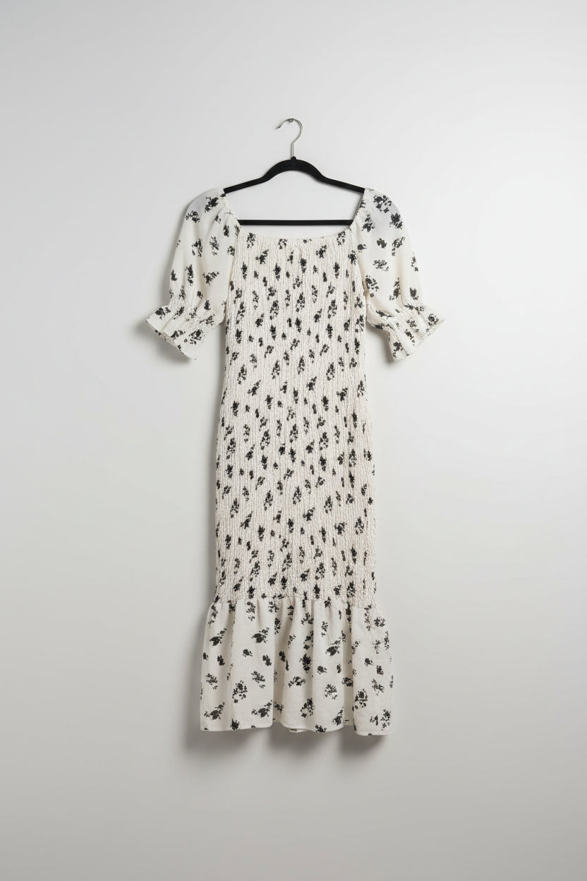 White dress with black floral pattern hanging on a wooden fence with greenery in the background