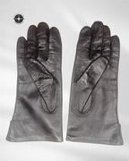 A pair of black leather gloves laid flat on a light gray background, featuring classic hand-stitched detailing and a compass logo in the top-left corner.