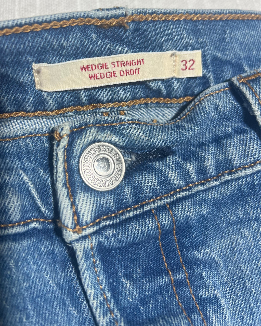 Close-up of the waistband of blue denim jeans, showing a cream-colored size label that reads 'WEDGIE STRAIGHT WEDGIE DROIT' and the size '32', along with a silver metal button.