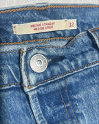 Close-up of the waistband of blue denim jeans, showing a cream-colored size label that reads 'WEDGIE STRAIGHT WEDGIE DROIT' and the size '32', along with a silver metal button.