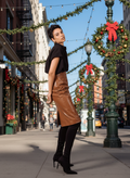 Woman in a black top and brown leather skirt standing on a city street decorated for Christmas.
