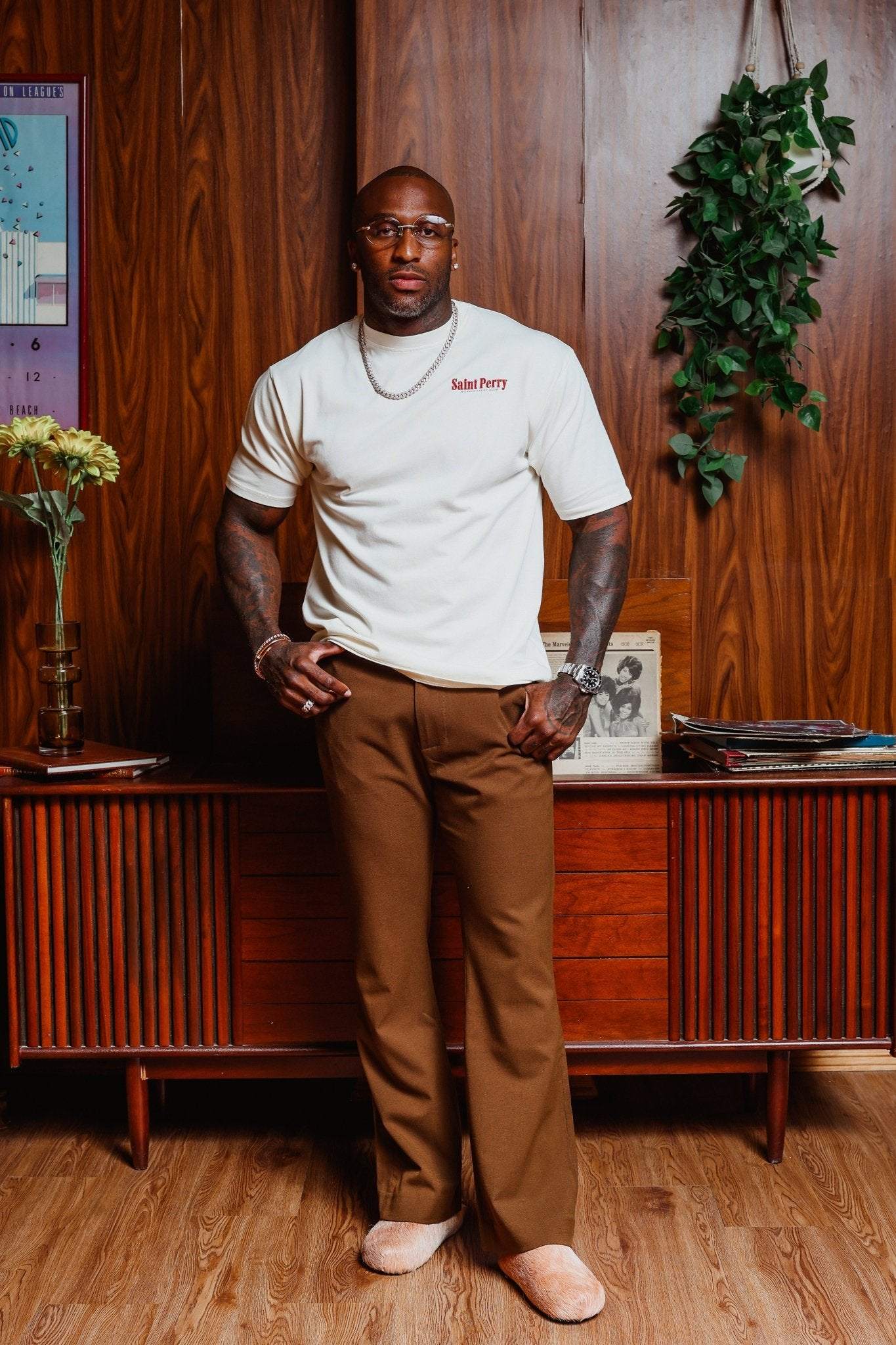 Man standing in a room with wooden walls and furniture, wearing a white t-shirt and brown pants.